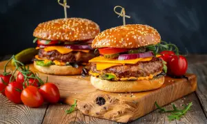 Dwa soczyste burgery z serem, warzywami i piklami na drewnianej desce, obok pomidorki koktajlowe.